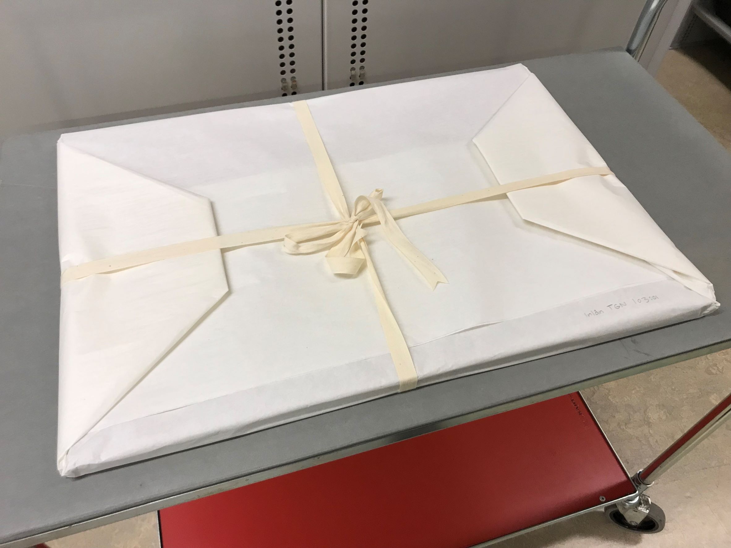 Ett platt vitt paket ombundet med vita bomullsband ligger på en transportvagn med hjul.