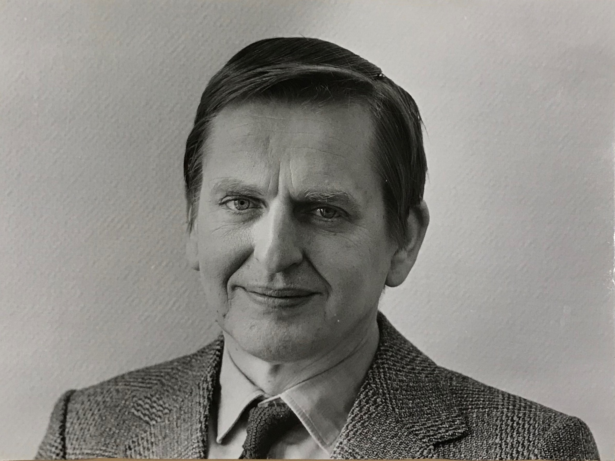 Svartvitt fotografi av en medelålders man som ler in i kameran, klädd i tweedkostym och en virkad slips.