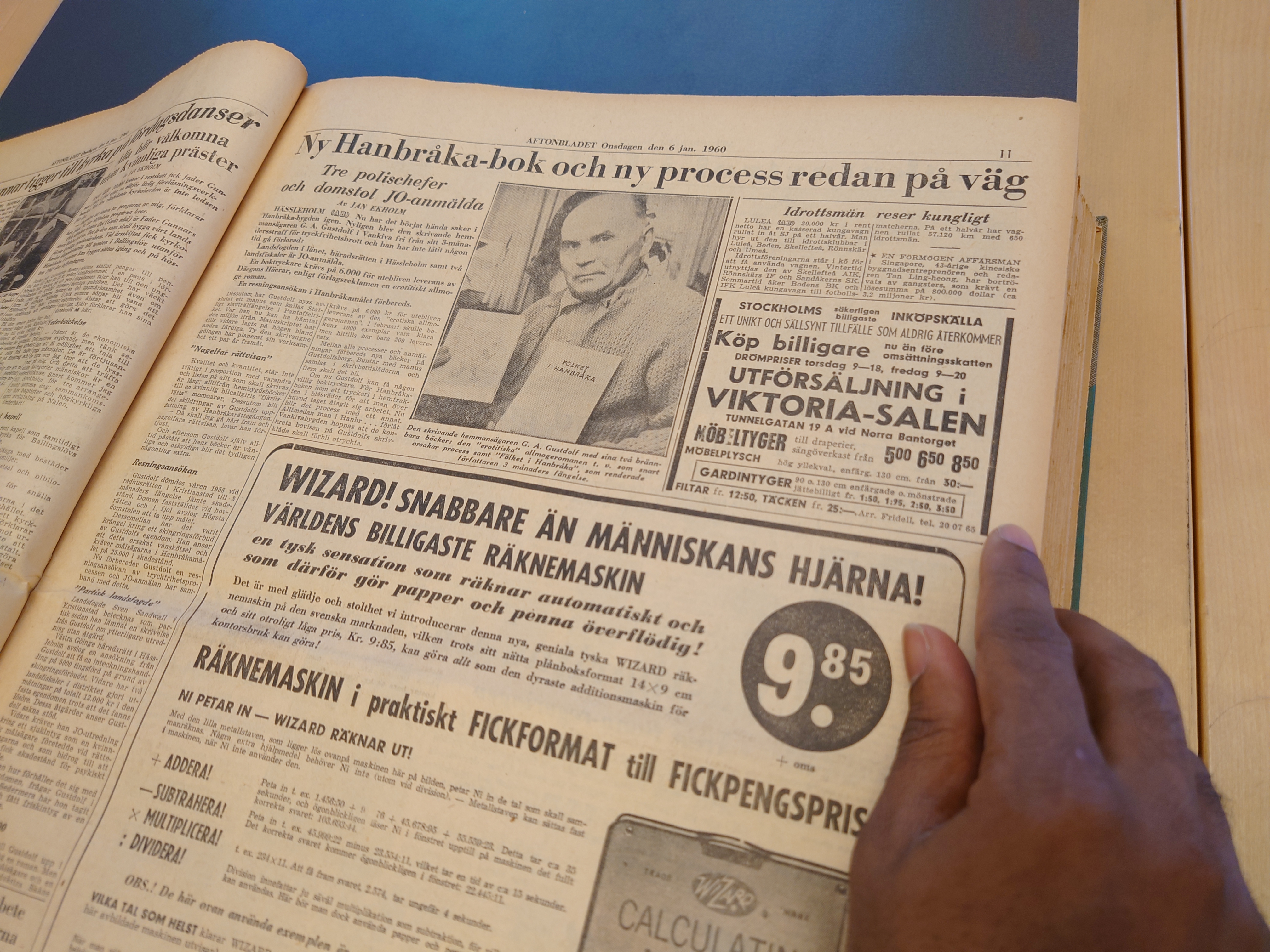 En hand bläddrar i ett tidningslägg med svartvit bild på medelålders man i stickad kofta, han håller i två böcker.