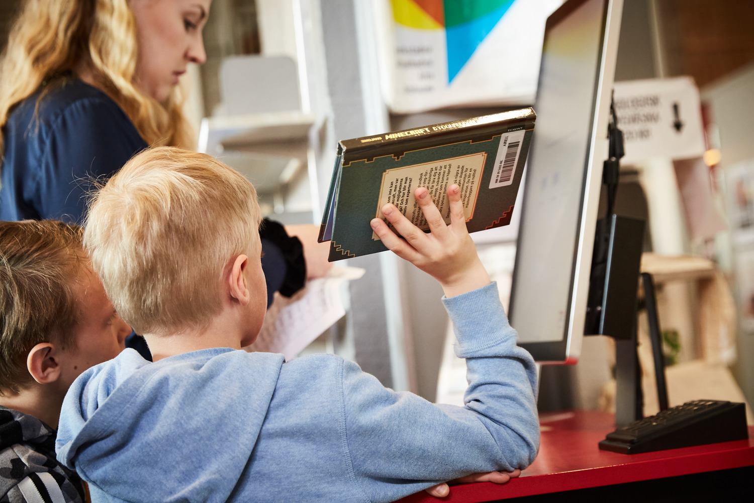 Ett barn håller en bok om Minecraft. Bredvid honom står ett annat barn och bakom dem en kvinna.