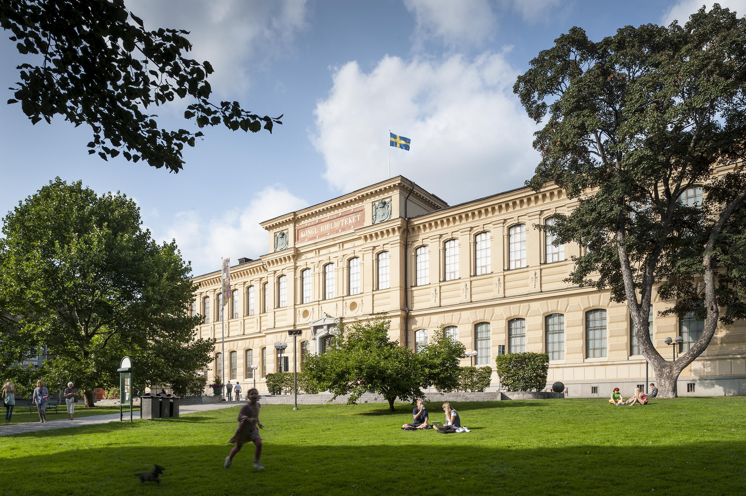 Gammal biblioteksbyggnad och gröna träd, blå himmel och vita moln i bakgrunden.