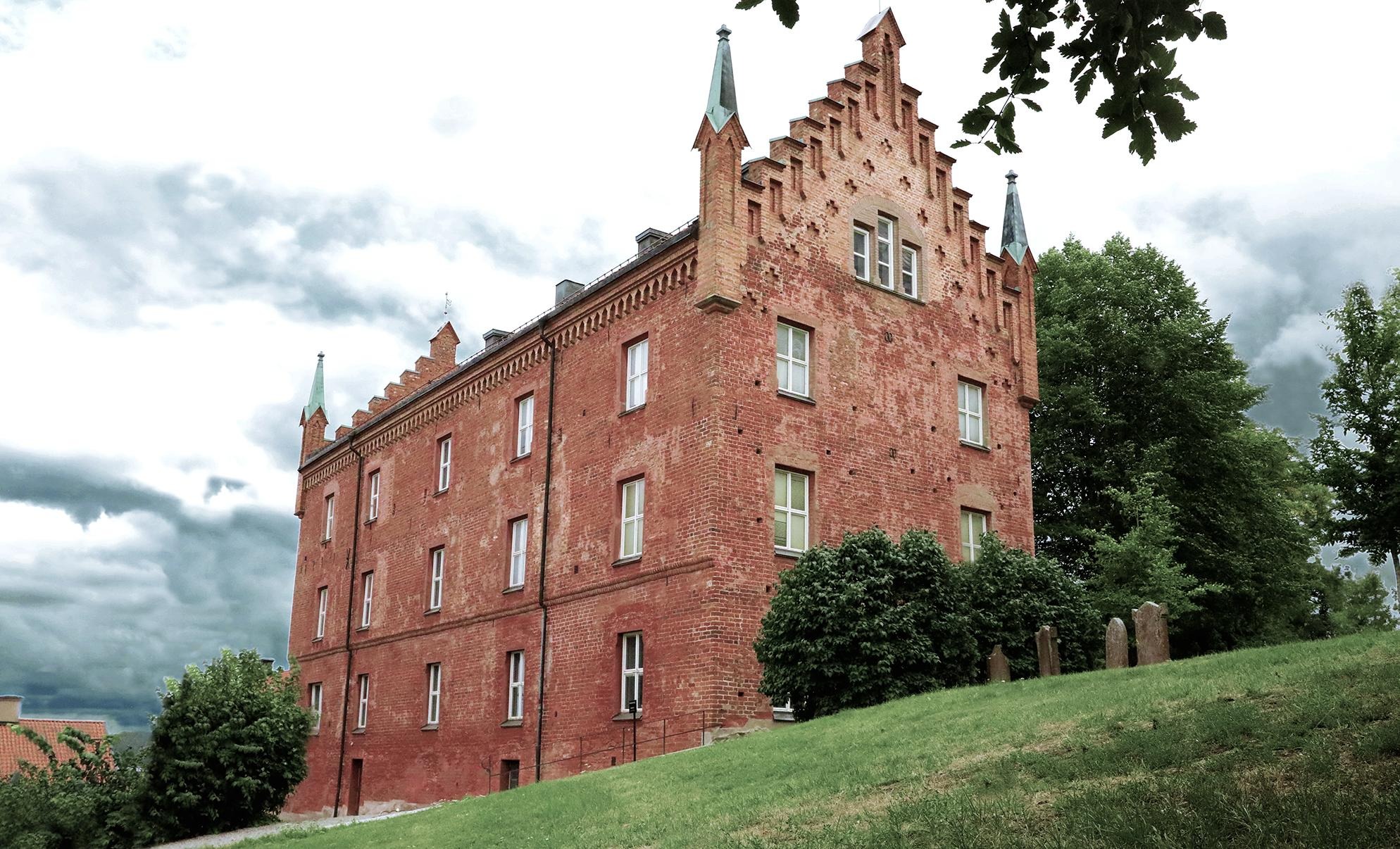 En byggnad i rött tegel mot en blå himmel med moln. Framför den syns en gräsmatta och gröna träd.
