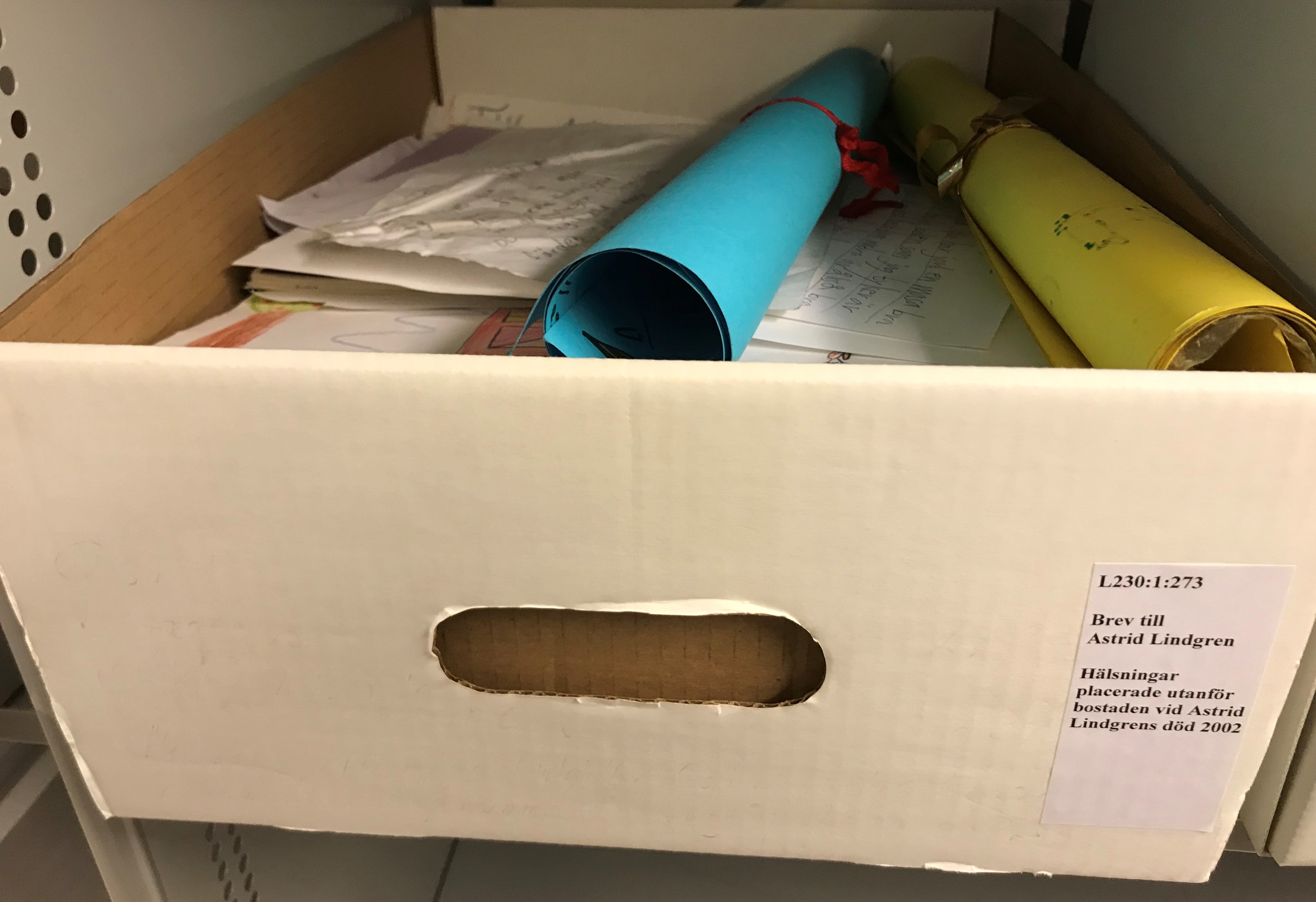 Stor vit kartong utan lock full med handskrivna papper och teckningar, överst ligger två färgglada rullar ombundna med snören.