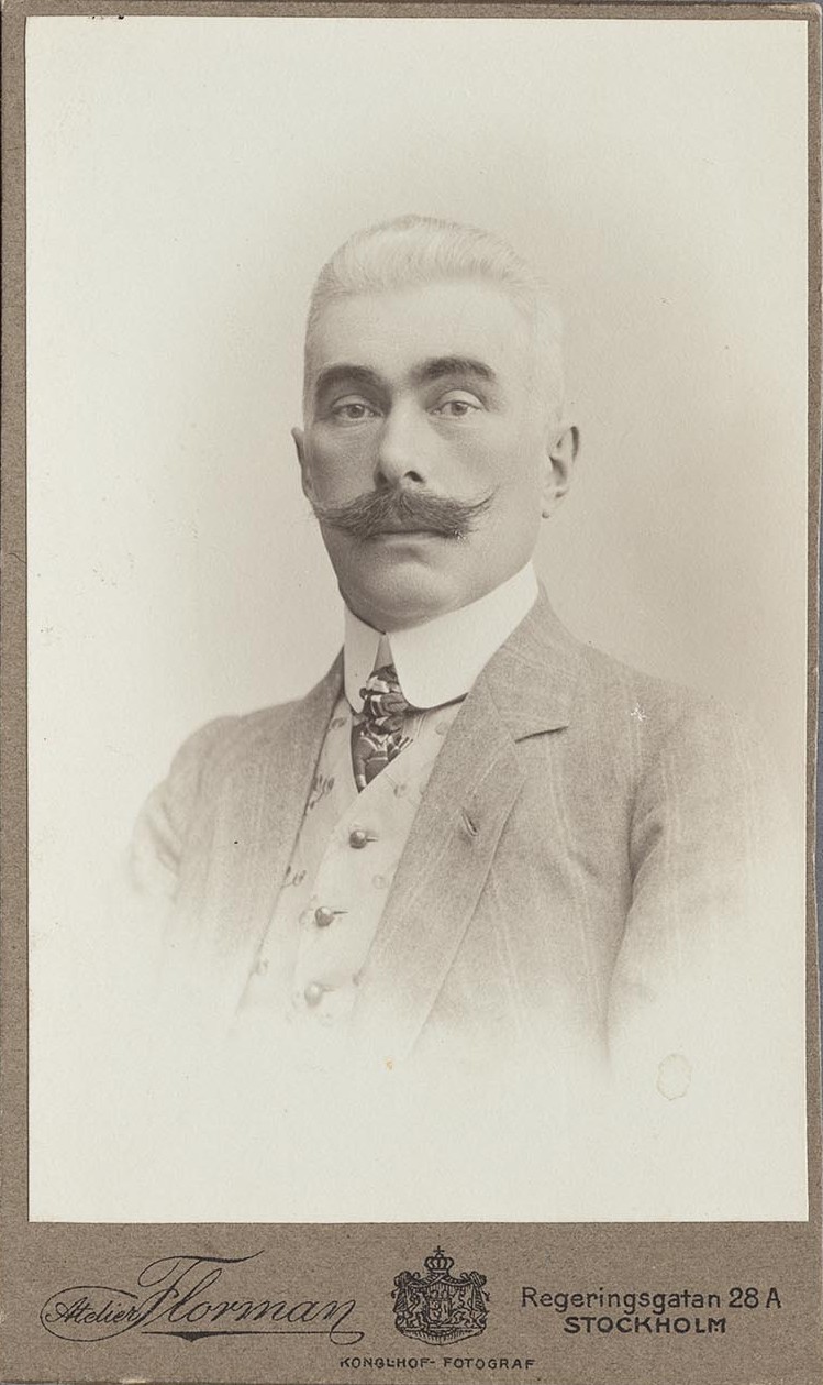 Svartvitt porträtt på välklädd man i mönstrad slips, kavaj och väst. Han har stora, bågformade ögonbryn och mustasch.