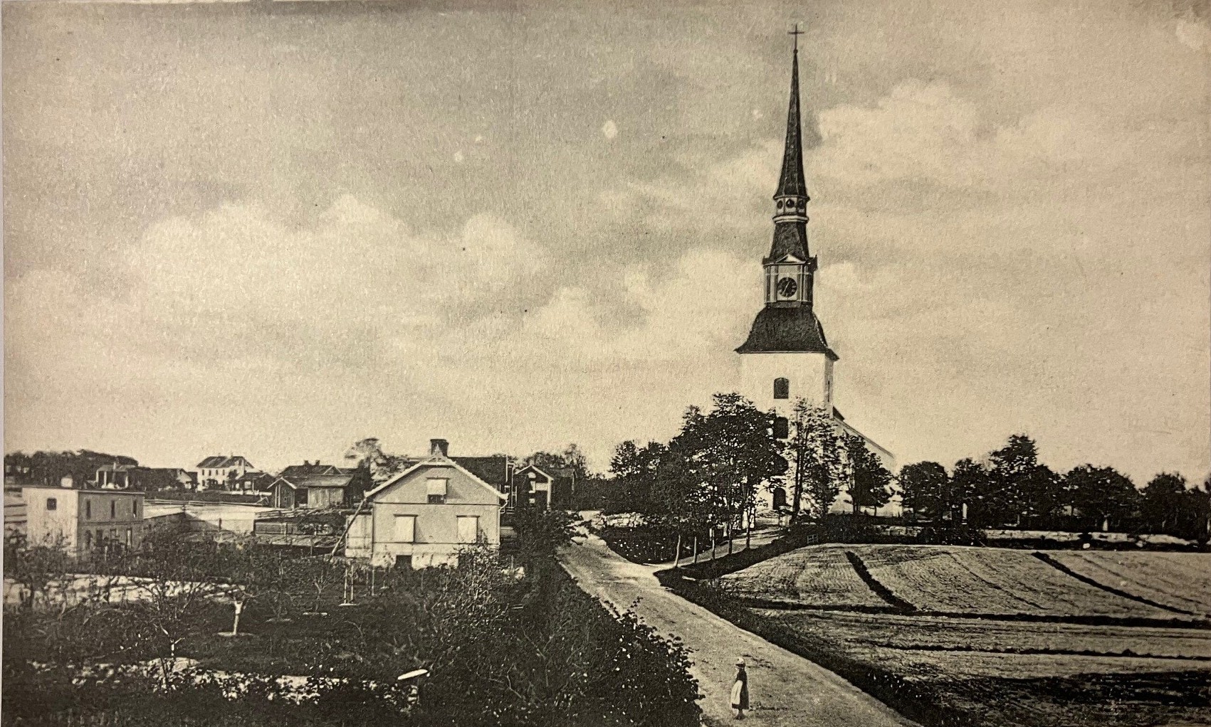 Svartvitt vykort med ett antal hus och en väg som leder fram till en kyrka.