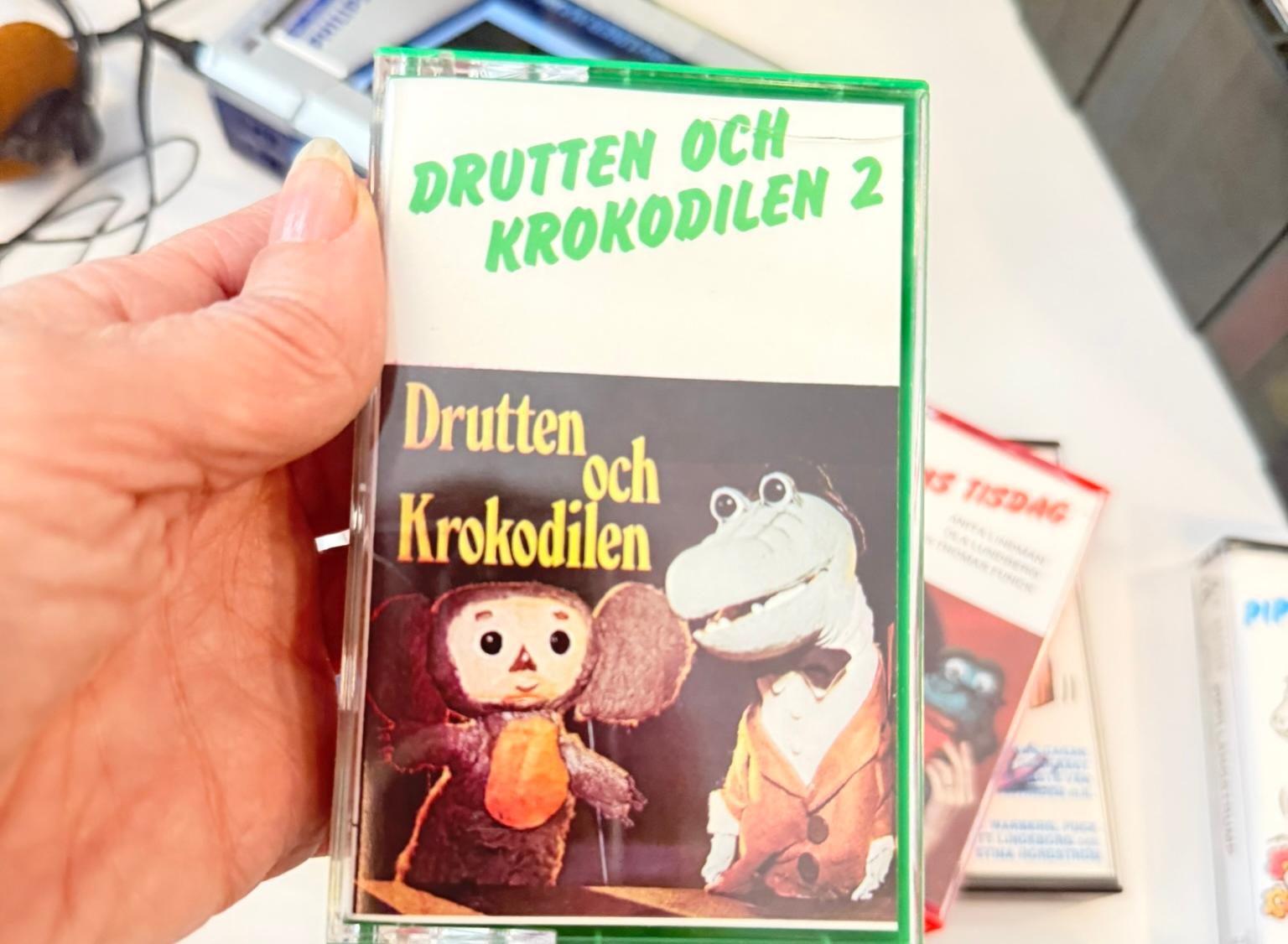 Hand håller upp en ljudkassett med bild på en brunt kramdjur med stora öron och en grön krokodil i hatt, fluga och kavaj.