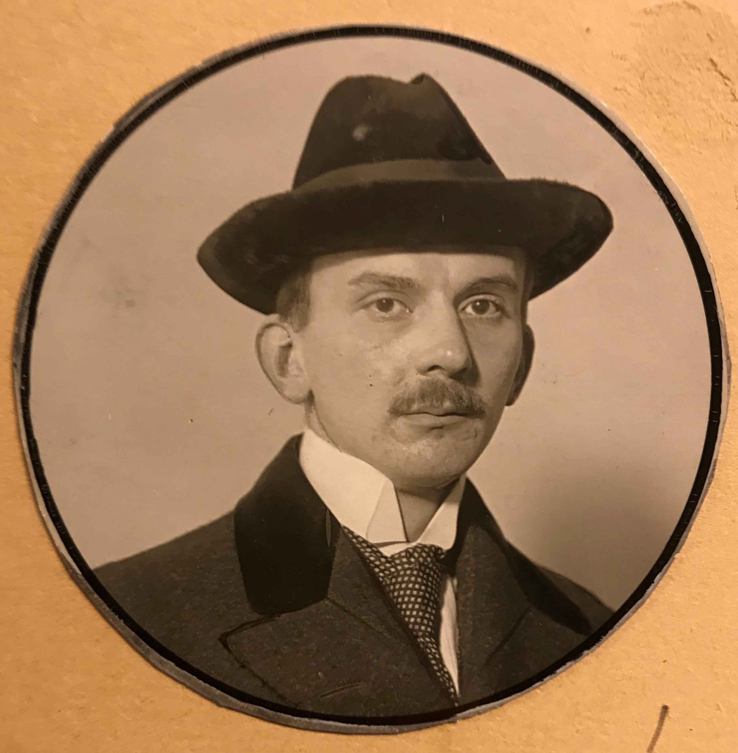 Cirkelformat fotografi av en ung man iförd hatt och mustasch.