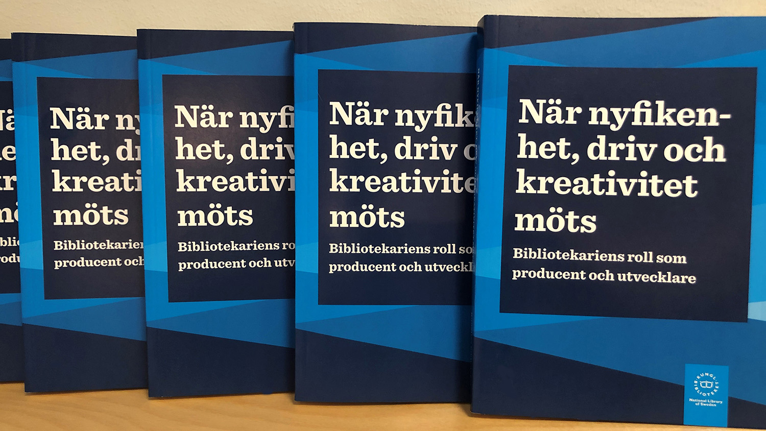En rad med blå böcker mot träyta. Text på omslaget: "När nyfikenhet, driv och kreativitet möts. Bibliotekariens roll som producent och utvecklare".