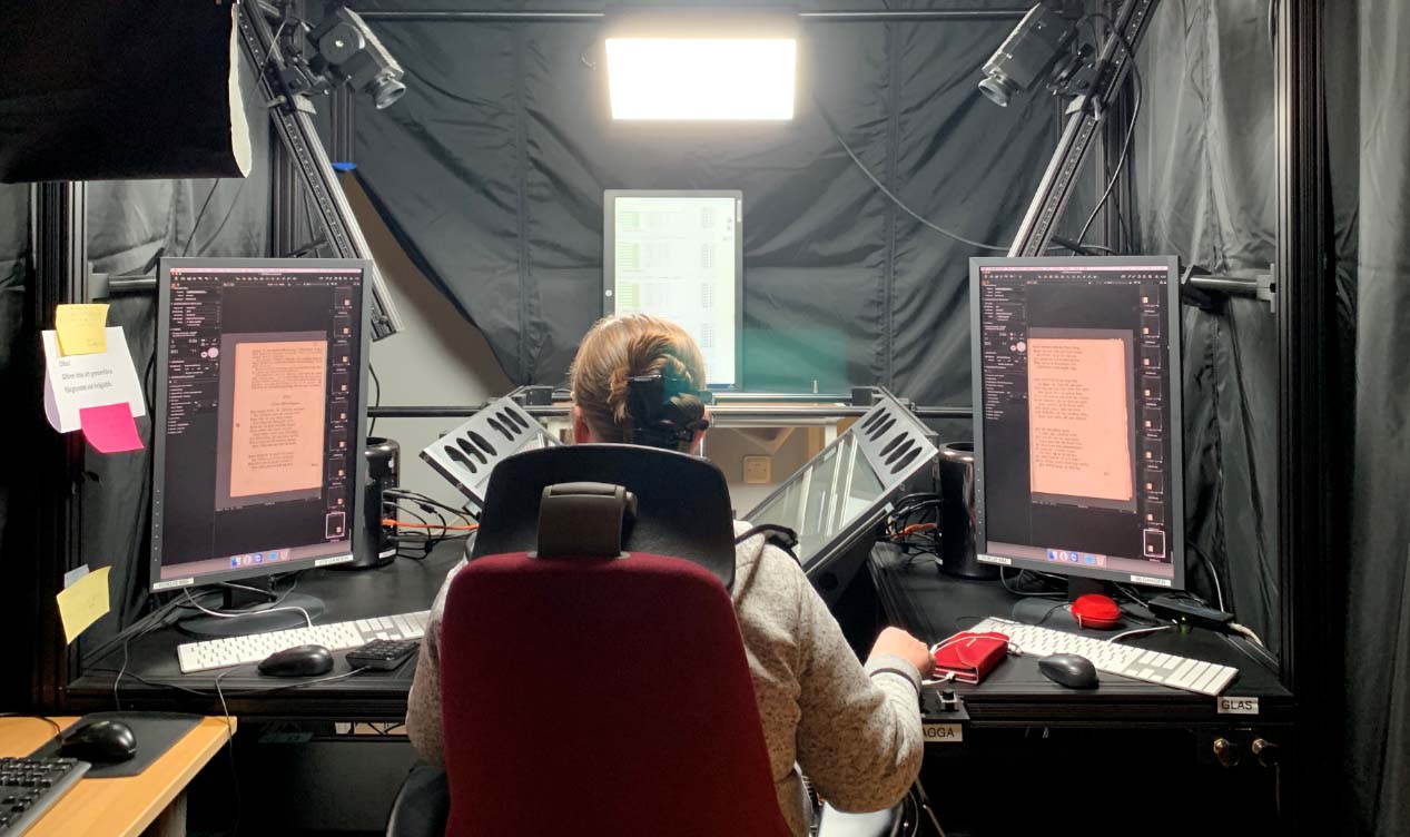 En person sitter framför utrustning för digitalisering på ett skrivbord. Bredvid utrustningen syns två skärmar som visar inskannade sidor av gammalt tryck.