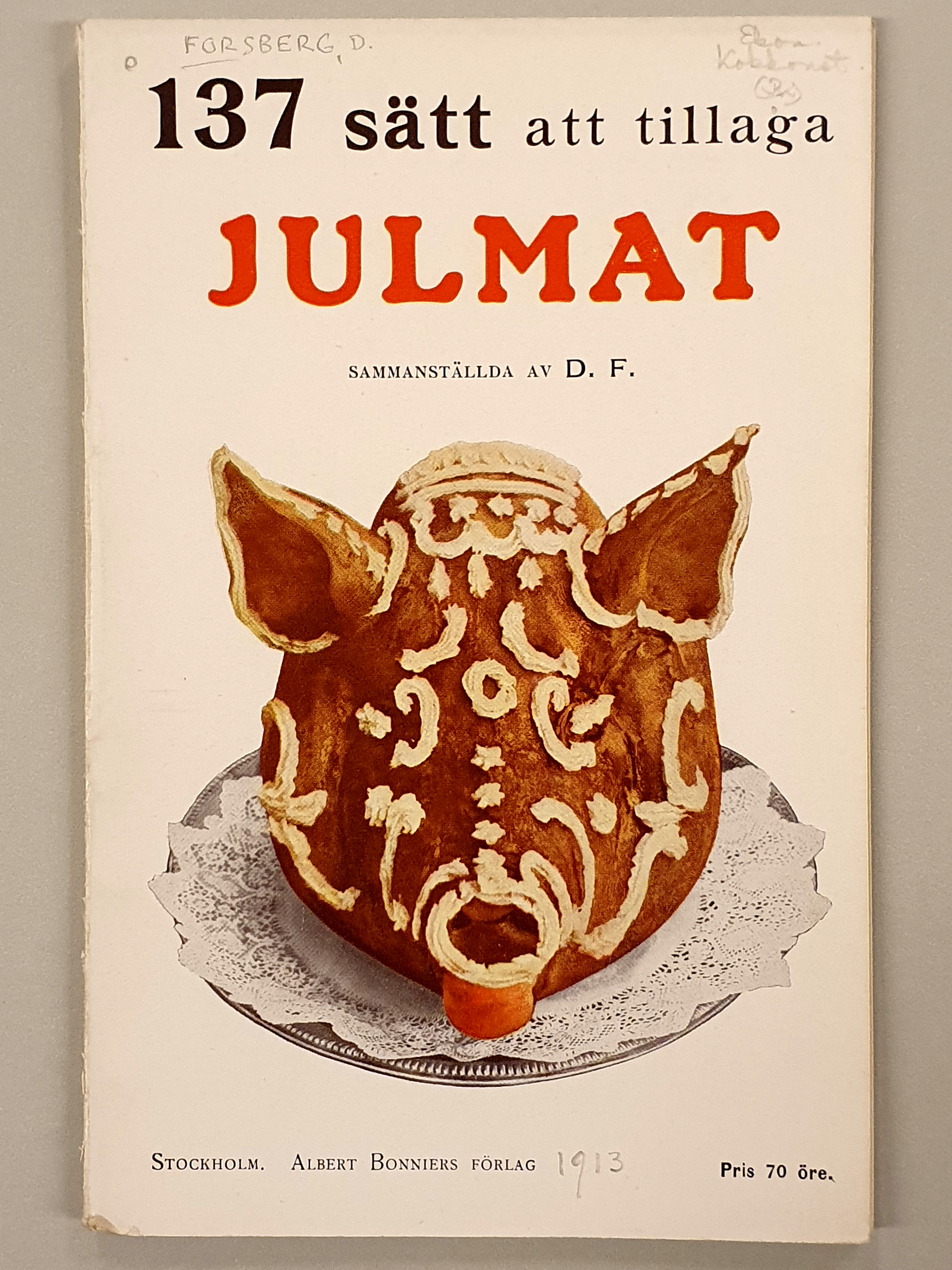Foto av en kokbok om julmat med svart och röd text mot en vit bakgrund. På omslaget finns även ett foto av ett grishuvud garnerat med kristyr.