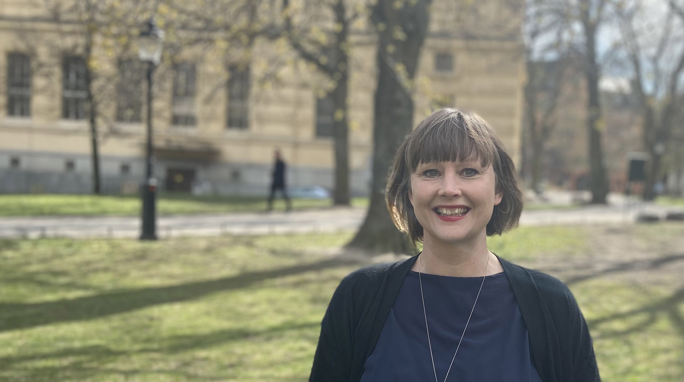 A smiling woman standing in a park in front of The National Library of Sweden, a yellow building.