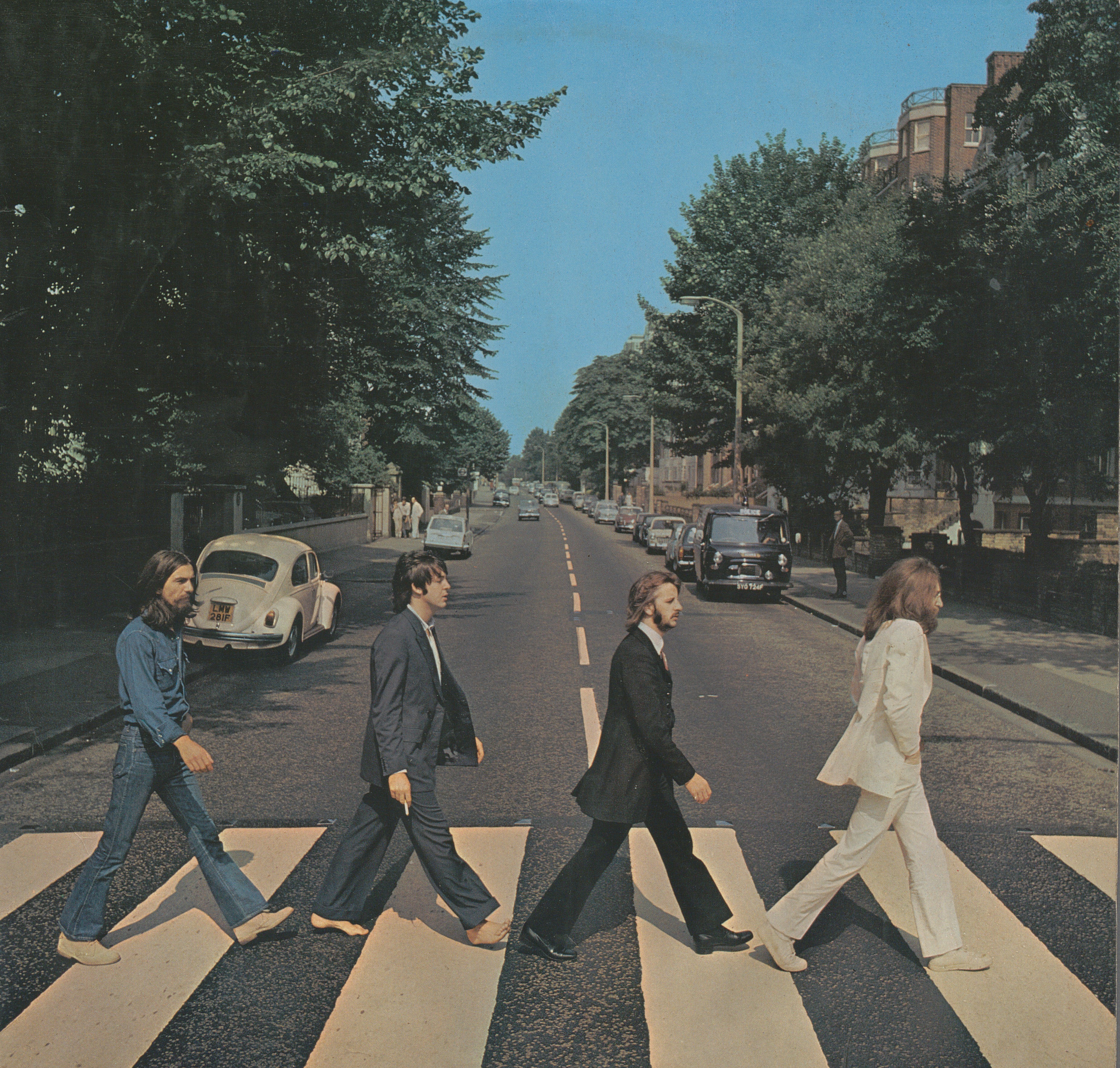 John Lennon, Ringo Starr, Paul McCartney och George Harrison går på ett övergångsställe. Paul barfota. Bilar står parkerade under grönskande träd.