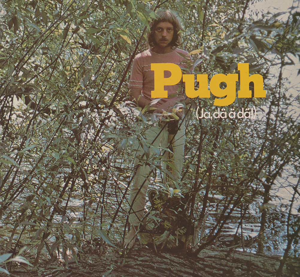 Pugh Rogefeldt står i rosa tröja delvis dold av trädgrenar med en gitarr framför sig vid vattnet. 