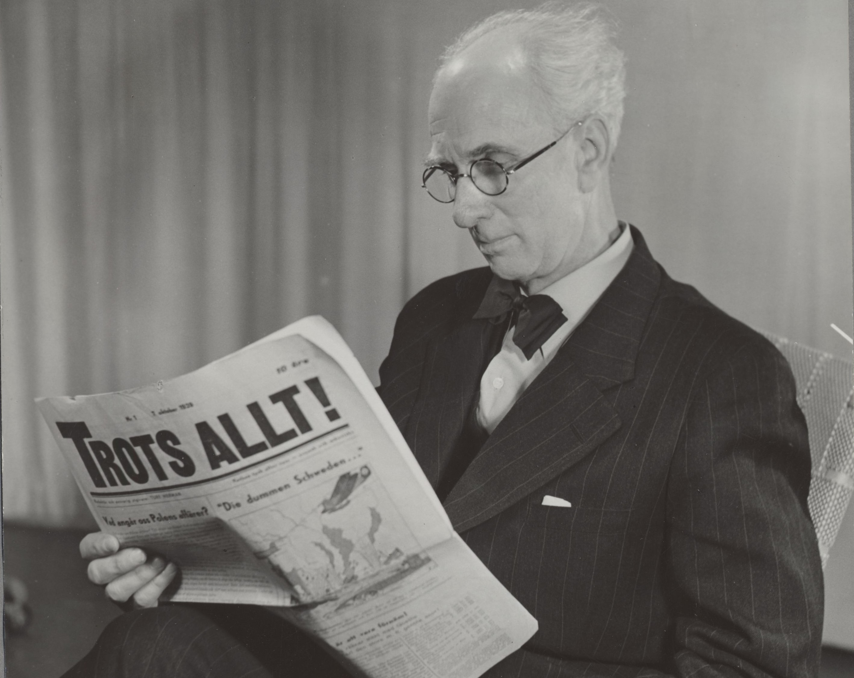 Svartvitt fotografi av en man med vitt hår och runda glasögon klädd i kostym som läser i en öppen tidning, på framsidan syns titeln "Trots allt"