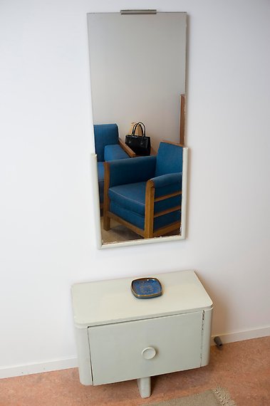 En avlång spegel hänger på väggen ovanför en liten byrå med ett blått fat. Spegelbilden visar en blå soffa.