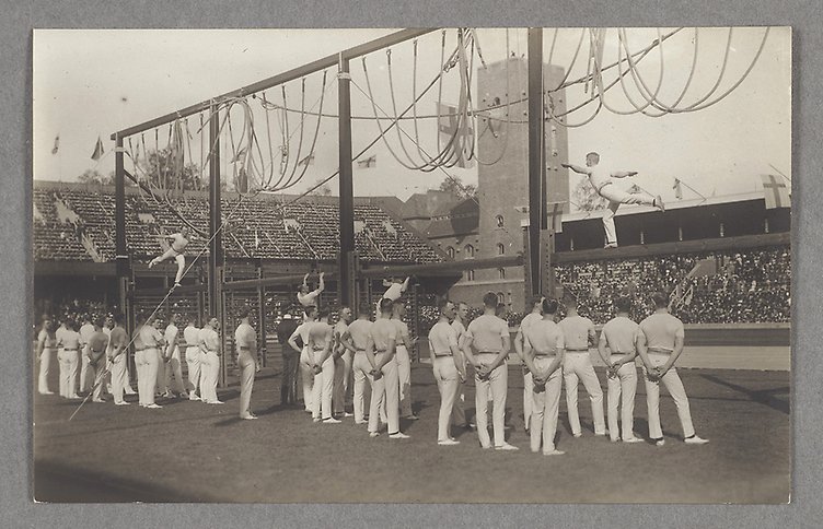 Fotografi av en grupp vitklädda manliga gymnaster vid en hög gymnastikställning på en arena full med publik. 