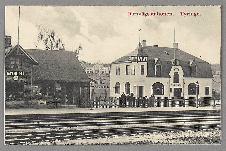 Svartvitt fotografi av ett stationshuset och en tågperrong med en grupp människor.