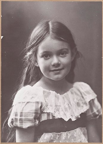 Svartvitt fotografi av en liten flicka med långt mörkt hår och rutig klänning.
