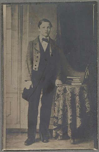 Svartvitt porträtt av en ung pojke i helfigur stående vid ett bord med en trave böcker.