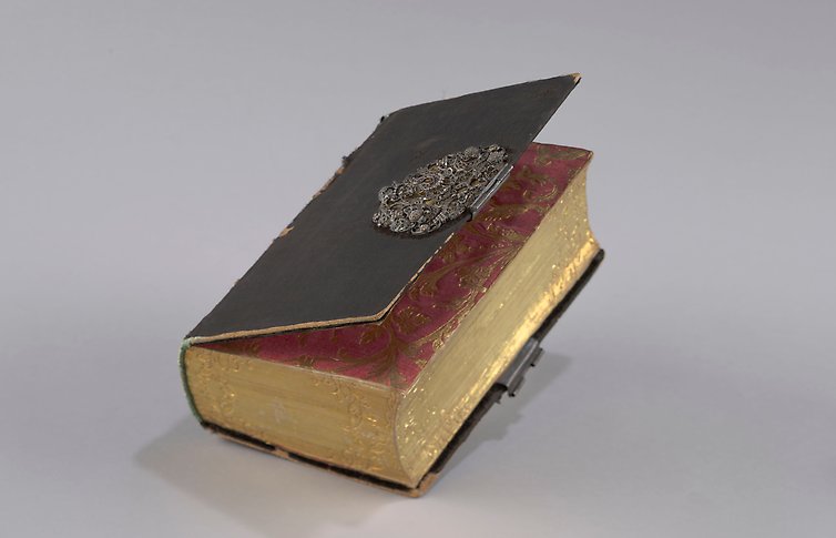 Svart äldre bok med utsmyckat metallspänne. Sidorna dekorerade i rött och guld.