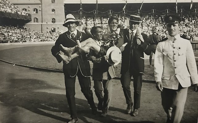 Svartvitt fotografi av en man med bara fötter och segerkrans som bärs av tre kostymklädda män inför fullsatta läktare på Stockholms stadion.