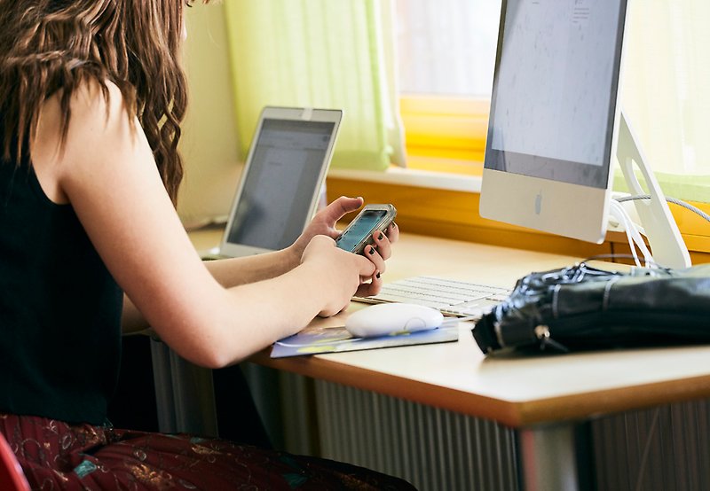 Kvinna sitter vid ett skrivbord framför en datorskärm med en telefon i händerna. Foto: Per & Per Fotograf AB