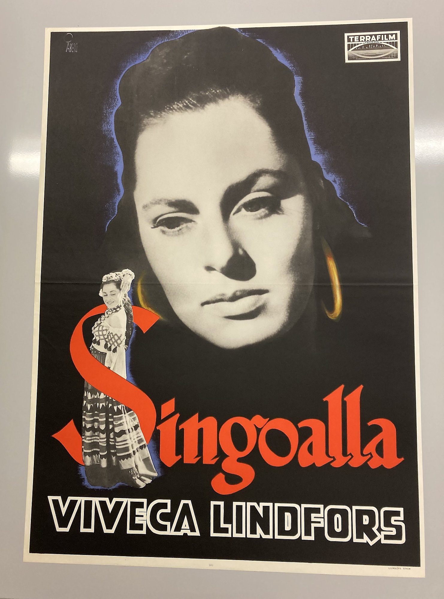 Färgfoto av en filmaffisch med en stor närbild på en vacker ung kvinna med stora ringar i öronen. Nertill en mindre svartvit bild av samma kvinna i helfigur iklädd romska kläder, stående inuti ett rött S som inleder titeln Singoalla.