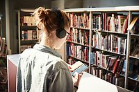 Foto av en person snett bakifrån, personen har på sig hörlurar, håller i och tittar på en smartphone i biblioteksmiljö