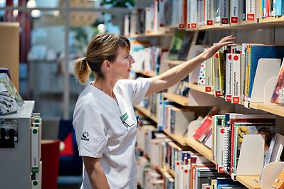 Kvinna i sjukhusdräkt tar en bok från en bokhylla.