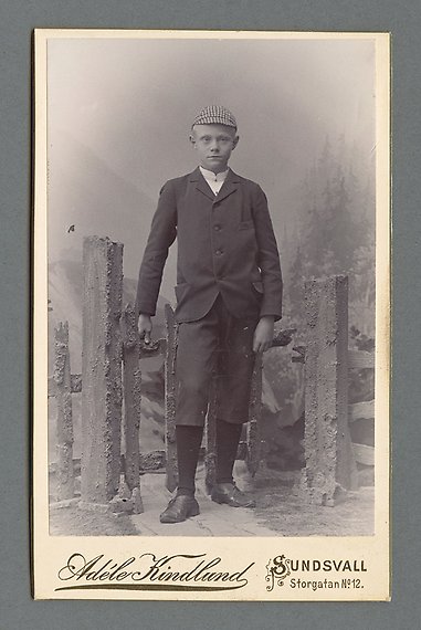 Svartvitt fotografi av pojke i mörk kostym med kortbyxor, höga strumpor och rutig keps. Han står vid en gammal grind och i bakgrunden skymtas skog och berg.
