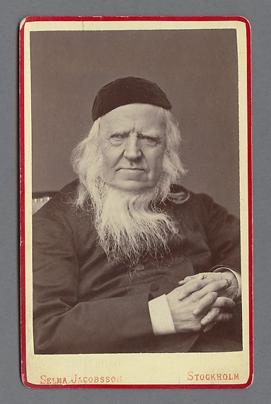 Svartvitt fotografi av äldre man med vitt långt hår och skägg. Han bär mörk kavaj och hätta på huvudet.