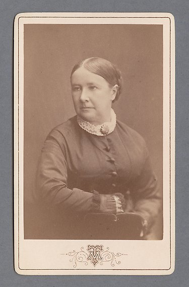 Svartvitt fotografi av kvinna i stramt uppsatt frisyr med knut och mittbena och klänning med brosch vid halsen. Hon tittar snett ut ur bild.