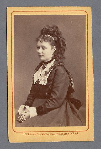 Svartvitt fotografi av kvinna med mörkt lockigt hår uppsatt i en hög frisyr. Hon sitter ner med händerna knäppta i knäet och ser ut ur bild.