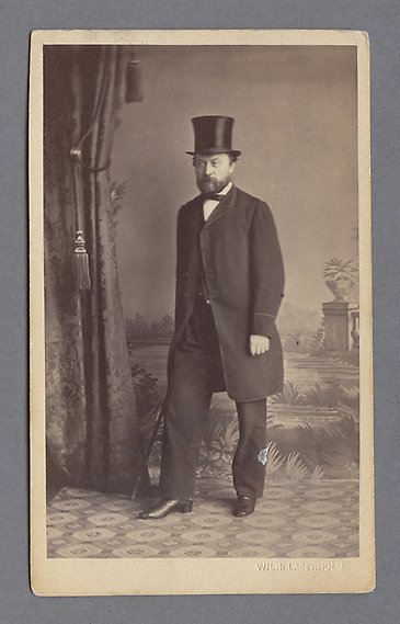 Svartvitt fotografi av herre i hög hatt, kostym och käpp står på ett mönstrat golv bredvid ett påkostat draperi.