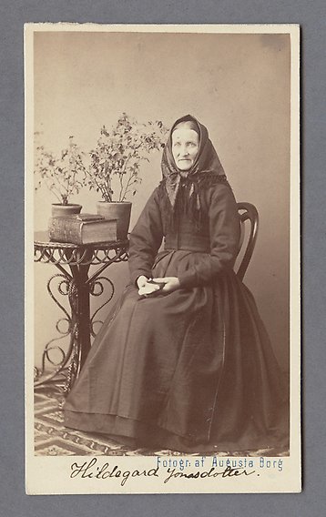 Svartvitt fotografi av kvinna i svart klänning och svart schal på huvudet. Hon sitter med en bok i knät bredvid ett litet bord med bok och krukväxter