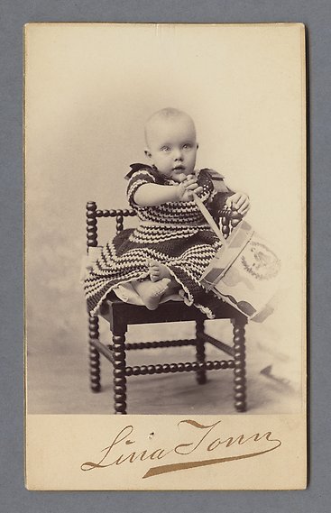 Litet barn i stickad klänning sitter på en svart trästol med en trumma och trumpinne.