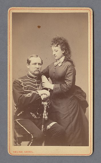 Svartvitt fotografi av man i militäruniform som sitter ner lutad mot en sabel. Intill står en kvinna lutad med armarna mot hans axel. 
