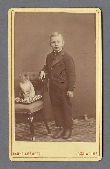Svartvitt fotografi av pojke i svart kostym som lutar armen mot en klädd stol där det sitter en katt. 