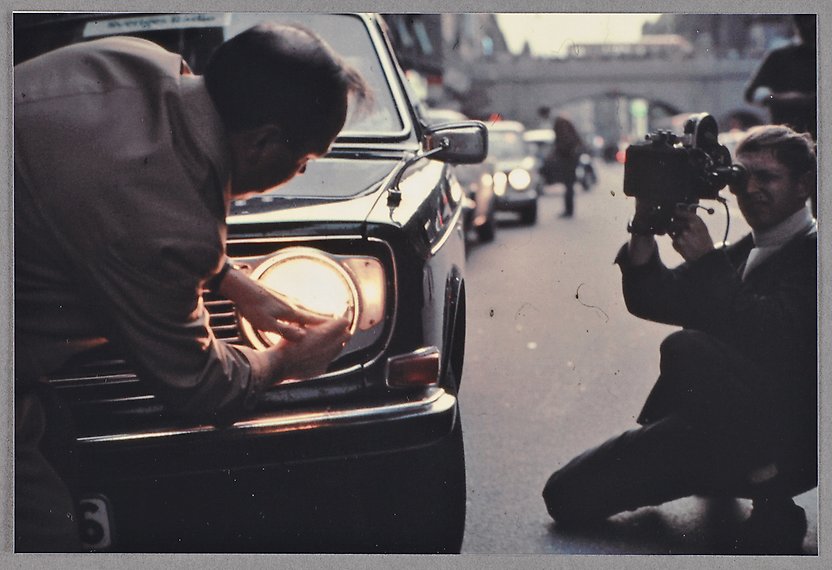 Färgfotografi av fronten på en bil där föraren justerar billamporna. En man sitter på knä och filmar.