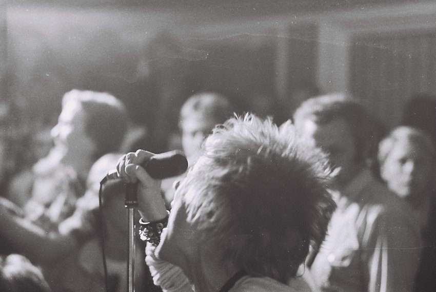 Svartvitt fotografi av en man med rufsigt blont hår som sjunger i mikrofon framför publik