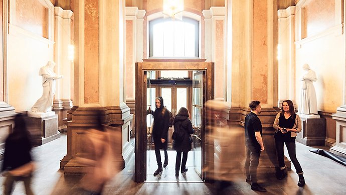 Entréhallen i Kungliga biblioteket, med höga, vita pelare och människor som pratar och passerar.