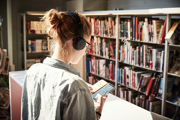 Foto av en person snett bakifrån, personen har på sig hörlurar, håller i och tittar på en smartphone i biblioteksmiljö